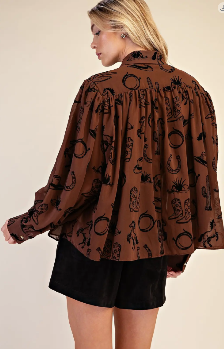 A woman shown from the back wearing a brown long-sleeve blouse with a black western-themed print of horseshoes, cacti, and boots, paired with black shorts, against a solid beige background.