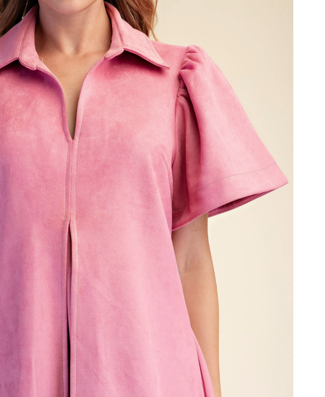 Close-up of a model wearing a pink suede-like short-sleeve collared top or dress with a front zipper and gathered puff sleeves, against a light beige background.