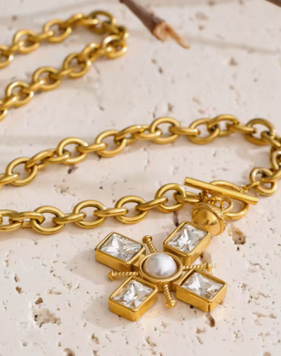 Gold-tone chain necklace with a cross-shaped pendant featuring clear square stones, a central white pearl, and a toggle clasp, displayed on a textured beige stone surface.