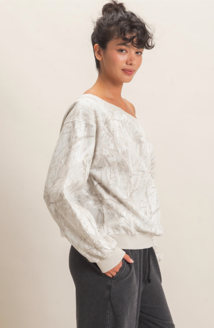 A woman wearing a textured, off-white long-sleeve knit sweater with a subtle abstract pattern, paired with dark gray sweatpants, photographed against a solid light beige background.