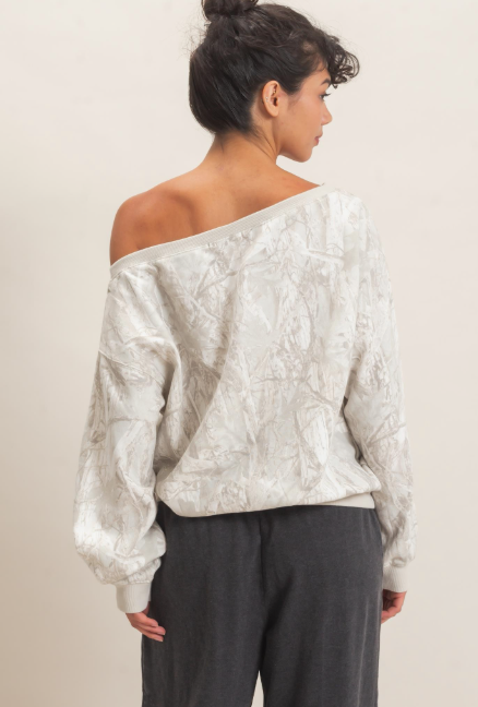 A woman shown from the back wearing a textured off-the-shoulder long-sleeve top in a light cream or off-white color, paired with dark gray pants, against a plain light beige background.