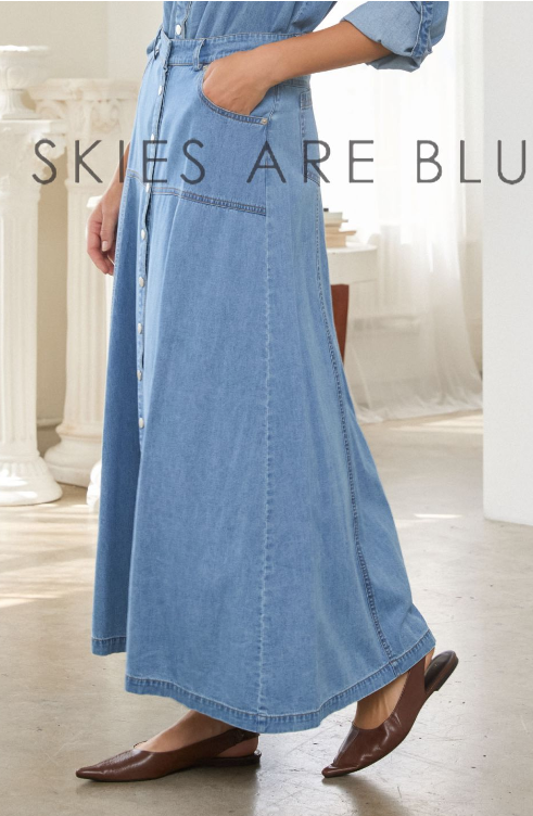 A person wearing a light blue denim maxi skirt with front pockets and a button front, paired with brown pointed flats, standing in a bright indoor room. Large text across the image reads 'SKIES ARE BLU'.