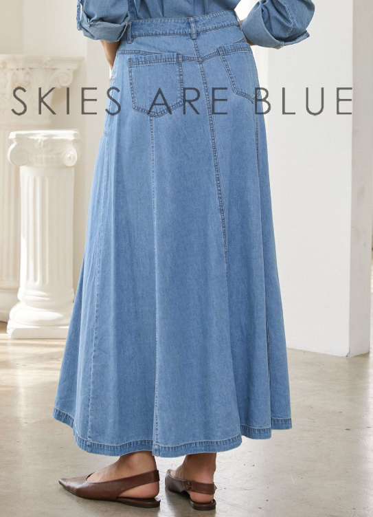 A model from the back wearing a light blue denim maxi skirt with a button fly and back pocket details, styled with a matching blue denim shirt and brown flat sandals. The text 'SKIES ARE BLUE' is overlaid on the image.