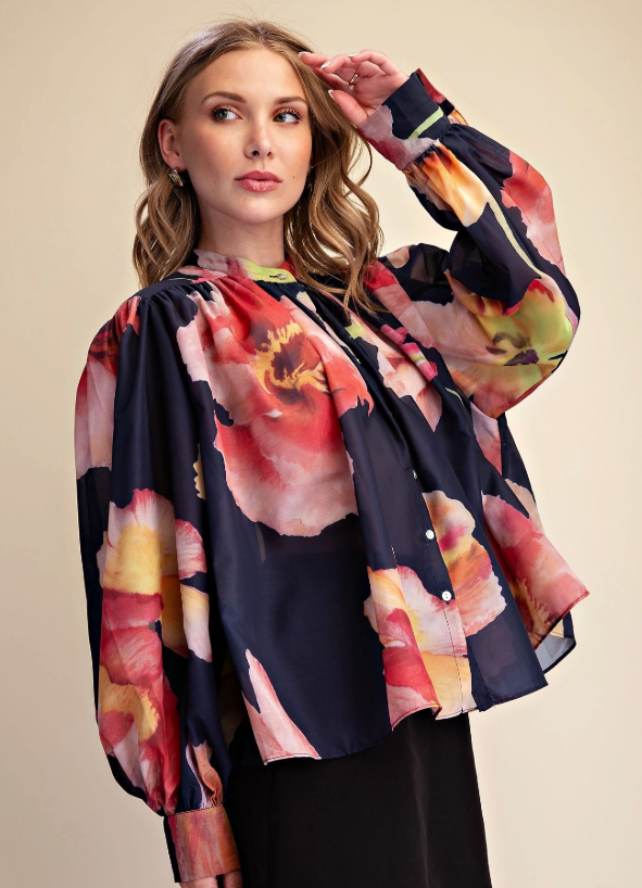 A woman wearing a long-sleeve button-front blouse with a bold floral print in red, pink, yellow, and orange over a dark navy base, posed against a plain beige studio background.