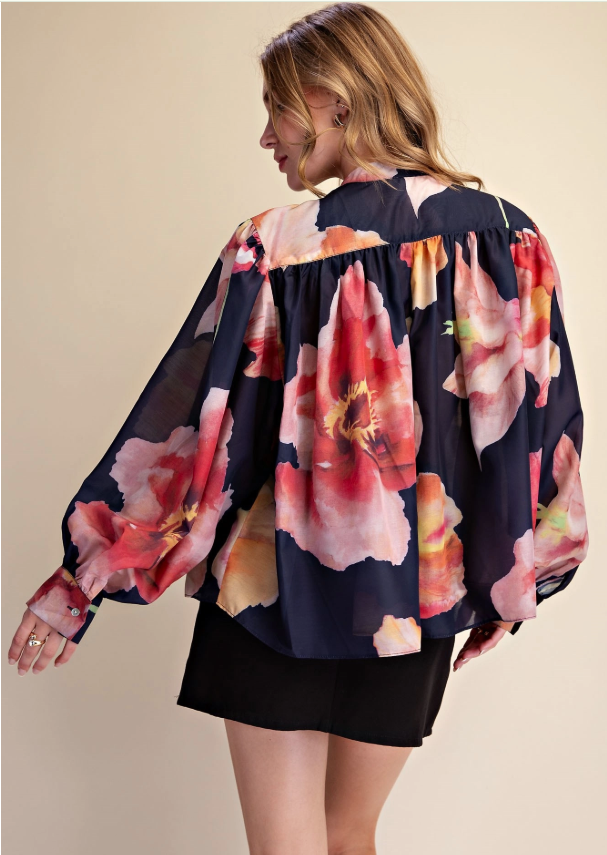 A woman wearing a navy blue blouse with a large pink and red floral print, paired with a black skirt, photographed against a solid beige background.