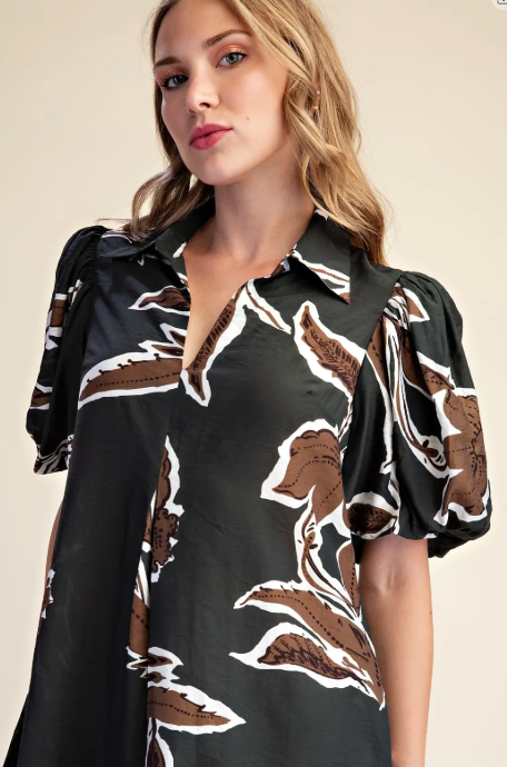 A woman wearing a black short-sleeve button-up shirt with a brown and white abstract leaf pattern, photographed against a plain beige studio background.