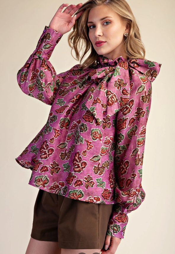 A woman modeling a long-sleeve blouse with a pink base and a multicolor floral and butterfly print, paired with brown shorts, against a solid beige background.