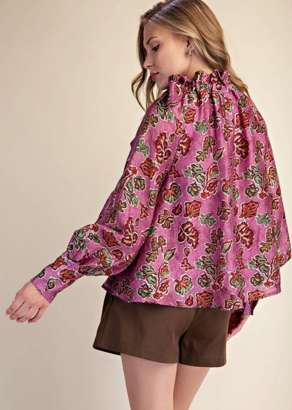 A woman wearing a pink long-sleeve blouse with a dense red, green, and gold floral print, paired with brown shorts, photographed against a solid beige studio background.
