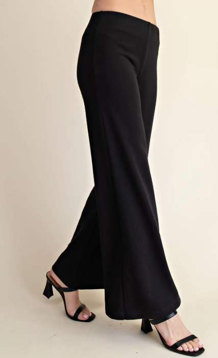 Black wide-leg pants shown on a model from the waist down, paired with black strappy heeled sandals against a plain beige studio background.