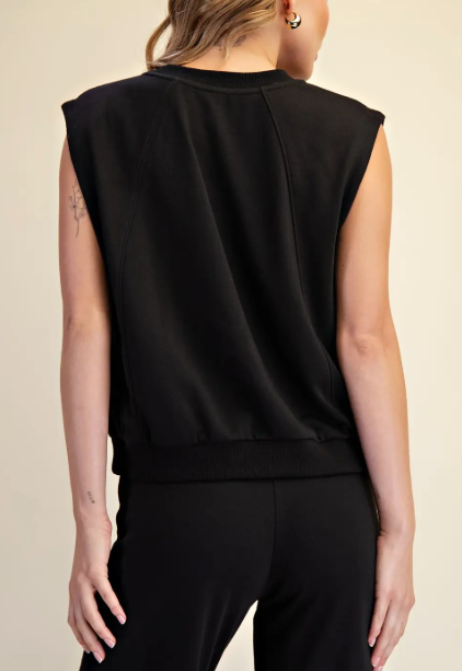 A woman shown from the back wearing a black sleeveless knit top with a crew neck and ribbed hem, paired with black pants, against a plain beige studio background.