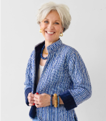 A smiling older woman with short white hair wearing a blue and white vertically striped jacket with dark navy cuffs and collar, photographed against a plain light gray background.