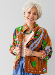 An older woman with short white hair smiling while wearing a brown fringed cardigan decorated with colorful floral and leaf patterns, holding a matching patterned tote bag against a light gray studio background.