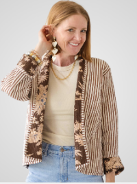 A woman wearing a brown and beige vertically striped open-front jacket with a floral pattern, layered over a cream top and blue jeans, photographed against a light gray studio background.