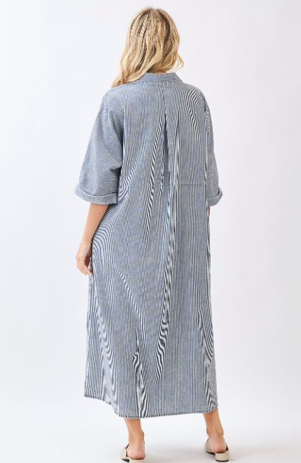 A woman viewed from the back wearing a long, loose button-front dress with vertical blue and white stripes, three-quarter sleeves, and rolled cuffs, photographed against a clean light gray studio background.