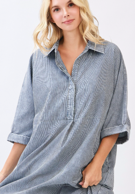 A woman wearing a loose, oversized button-front shirt dress with vertical blue and white stripes, photographed against a clean light gray studio background.