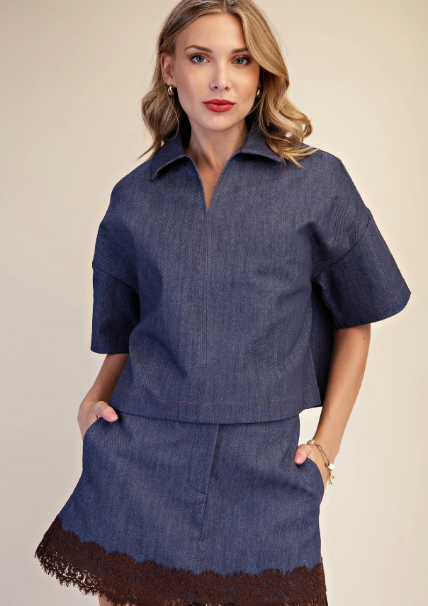 A woman modeling a dark blue denim two-piece set consisting of a short-sleeve collared top and a matching mini skirt with a brown lace hem, set against a plain beige background.