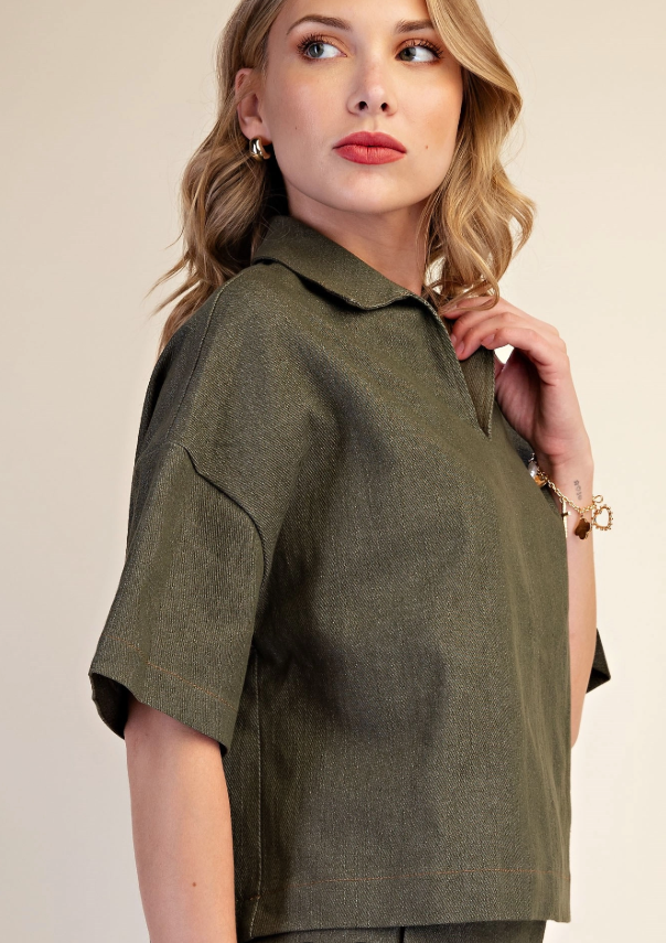 A woman wearing a short-sleeve olive green collared shirt, photographed against a plain light beige studio background.