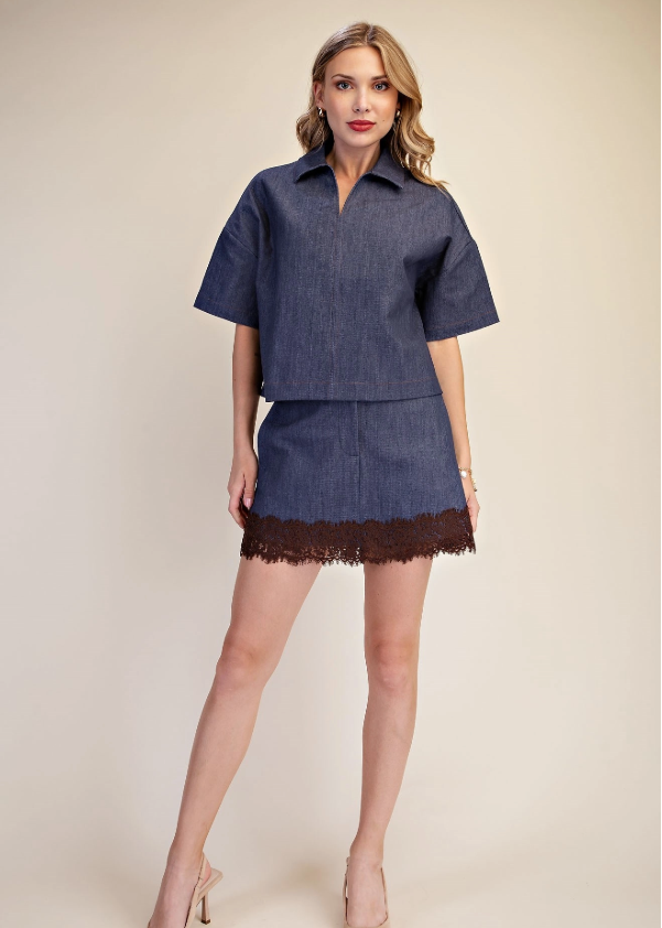 A woman models a two-piece denim set consisting of a short-sleeve button-up shirt and a matching mini skirt with a dark brown lace hem, set against a plain beige studio background.