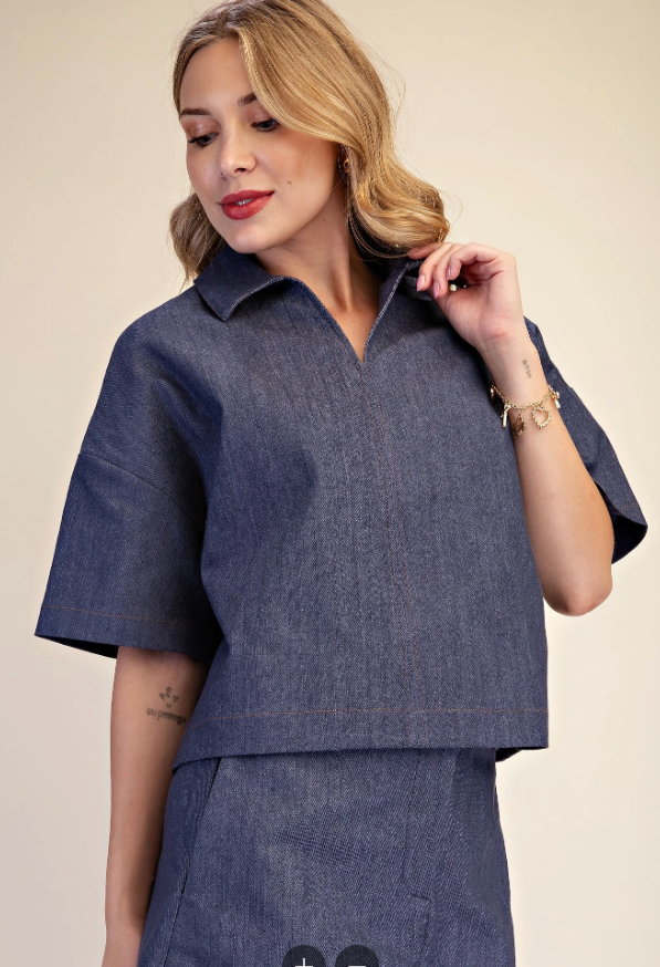 A woman wearing a dark blue denim-like short-sleeve collared top and matching skirt against a plain beige studio background.
