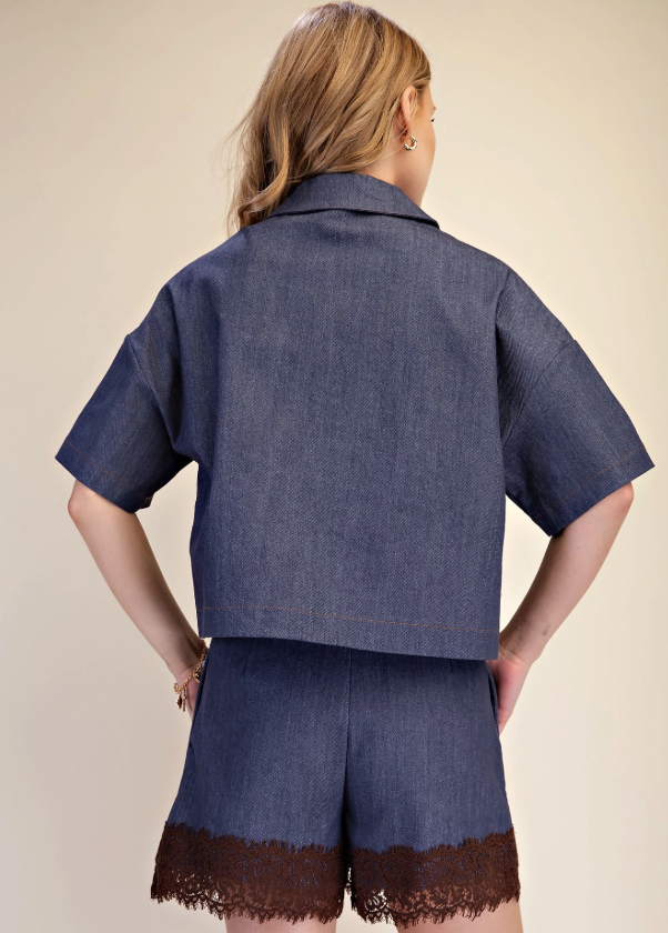 A woman shown from the back wearing a short-sleeve cropped denim jacket and matching denim shorts with a dark brown lace hem, against a plain beige studio background.