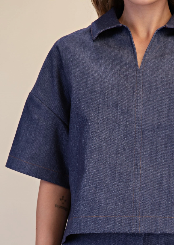 A close-up of a person wearing a cropped short-sleeve collared shirt in dark indigo denim with visible contrast stitching, against a plain light beige background.