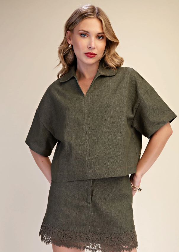 A woman modeling a matching olive green short-sleeve collared top and high-waisted mini skirt set with a lace hem, against a solid beige studio background.