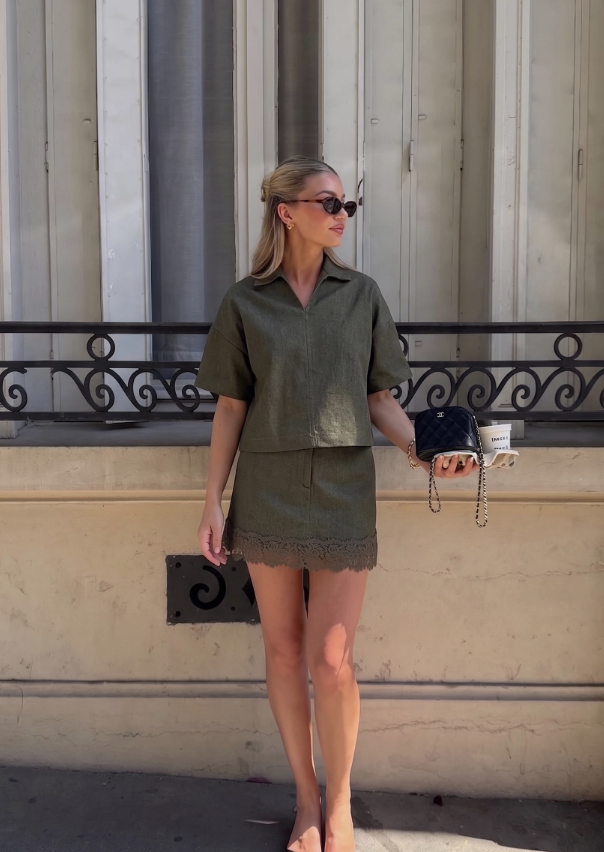 A woman stands outdoors against a beige building wall wearing a matching olive green short-sleeve collared top and skirt set, holding a small black handbag.