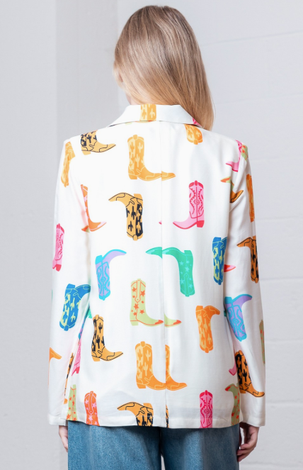 A woman viewed from behind wearing a white button-up shirt covered in a colorful cowboy boot print, paired with blue jeans against a white brick wall.
