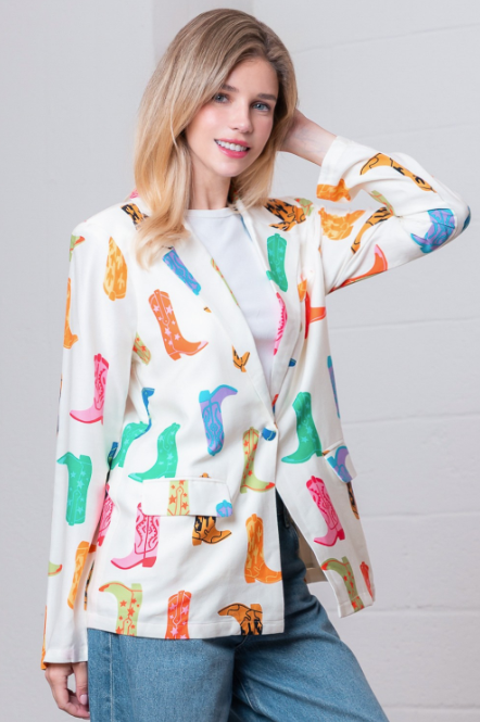 A woman wearing a white blazer covered in a colorful cowboy boot print, paired with a white top and blue jeans, posing against a white brick wall.