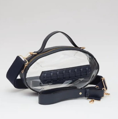A clear oval shoulder bag with navy blue trim, handle, and adjustable strap, featuring gold-tone hardware and a navy woven detail on the front, set against a light gray background.