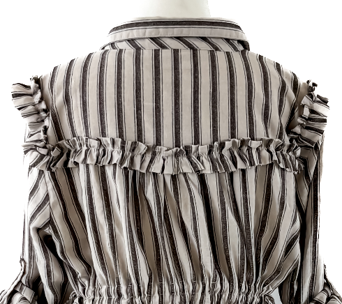 A close-up back view of a striped blouse displayed on a mannequin. The garment features vertical stripes in beige, brown, and black, with ruffled detailing along the shoulders and a gathered waist.