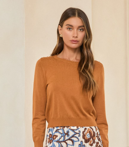 A woman wearing a ribbed knit crewneck sweater in a warm camel brown color, paired with a patterned skirt, standing against a neutral beige background.