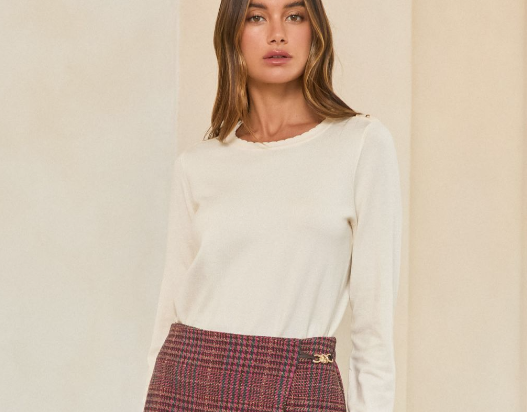 A woman wearing a cream-colored long-sleeve knit top and a red and blue plaid skirt, standing against a neutral beige wall.