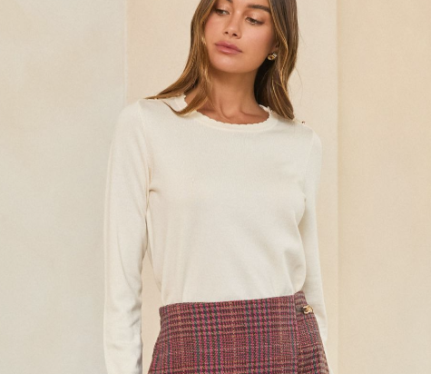 A woman wearing a cream-colored long-sleeve knit sweater and a red and blue plaid skirt, standing against a neutral beige wall.