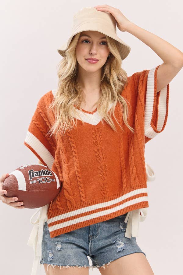 Woman wearing an orange sweater with white trim, holding a football, and posing against a plain background.