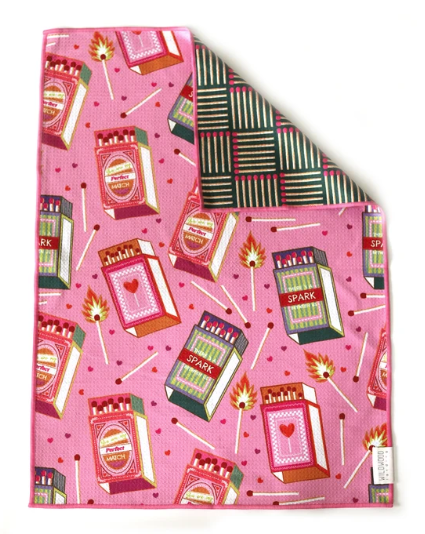 A square pink fabric item, likely a scarf or pocket square, featuring a repeating pattern of matchboxes, matchsticks, and small red hearts. The top-right corner is folded over to show the reverse side, which has a black, green, and pink striped pattern.