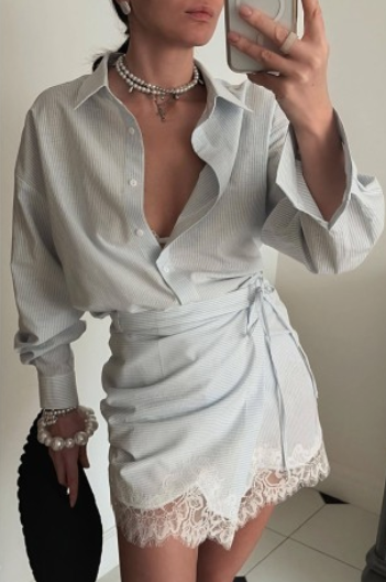 A woman takes a mirror selfie wearing a light gray and white vertically striped button-down shirt dress with a wrap tie waist and a lace hem. She is accessorized with layered pearl necklaces and bracelets, holding a black clutch.