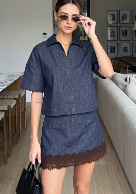 A woman models a matching two-piece denim outfit consisting of a short-sleeve collared top and a mini skirt with a brown lace hem, styled with sunglasses and a black handbag in a modern living room setting.