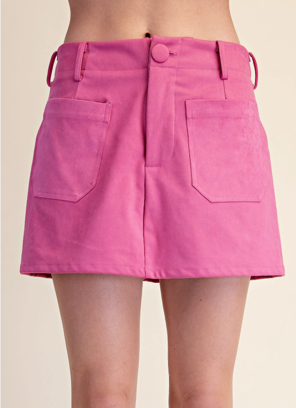 A close-up of a model wearing a bright pink mini skirt with front patch pockets and a button closure, shown against a plain beige background.