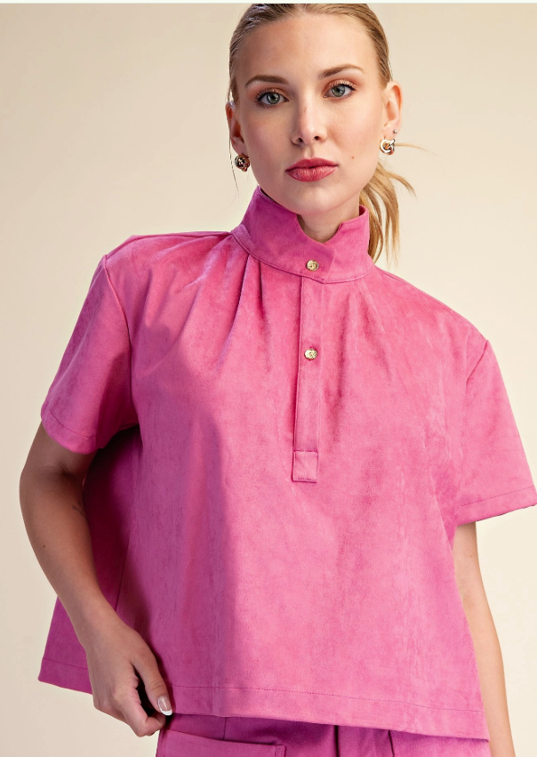 A woman modeling a bright pink short-sleeve top with a high mock neck and gold buttons, paired with matching pink bottoms, against a solid beige background.