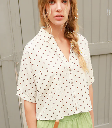 Woman wearing a white blouse with brown polka dots and green shorts against a wooden background