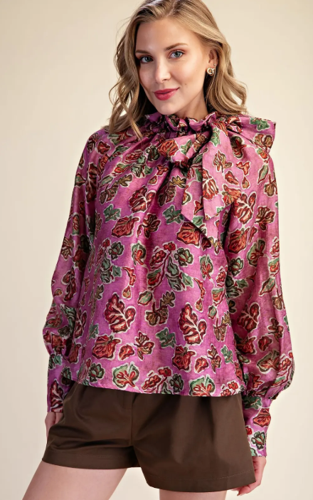 A woman wearing a pink long-sleeve blouse with a large ruffled bow at the neck and an allover floral print in red, green, and brown, paired with brown shorts, against a plain beige studio background.