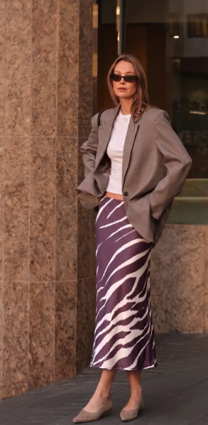 A woman stands outdoors against a stone column wearing an oversized taupe blazer, white top, and a purple-and-white zebra-print midi skirt, with nude pointed flats.