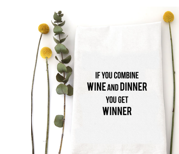 A white kitchen towel with black text reading 'IF YOU COMBINE WINE AND DINNER YOU GET WINNER' styled flat-lay on a white background with decorative eucalyptus stems and yellow flowers.