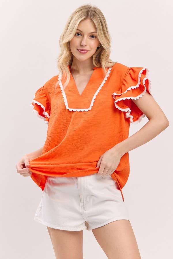 Woman wearing an orange top with ruffled details and white shorts on a light background