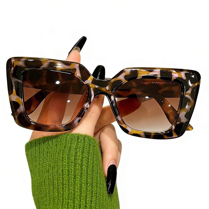 A hand wearing a green knit sweater holds a pair of oversized square sunglasses with a brown and black tortoiseshell patterned frame and gradient brown lenses against a plain white background.