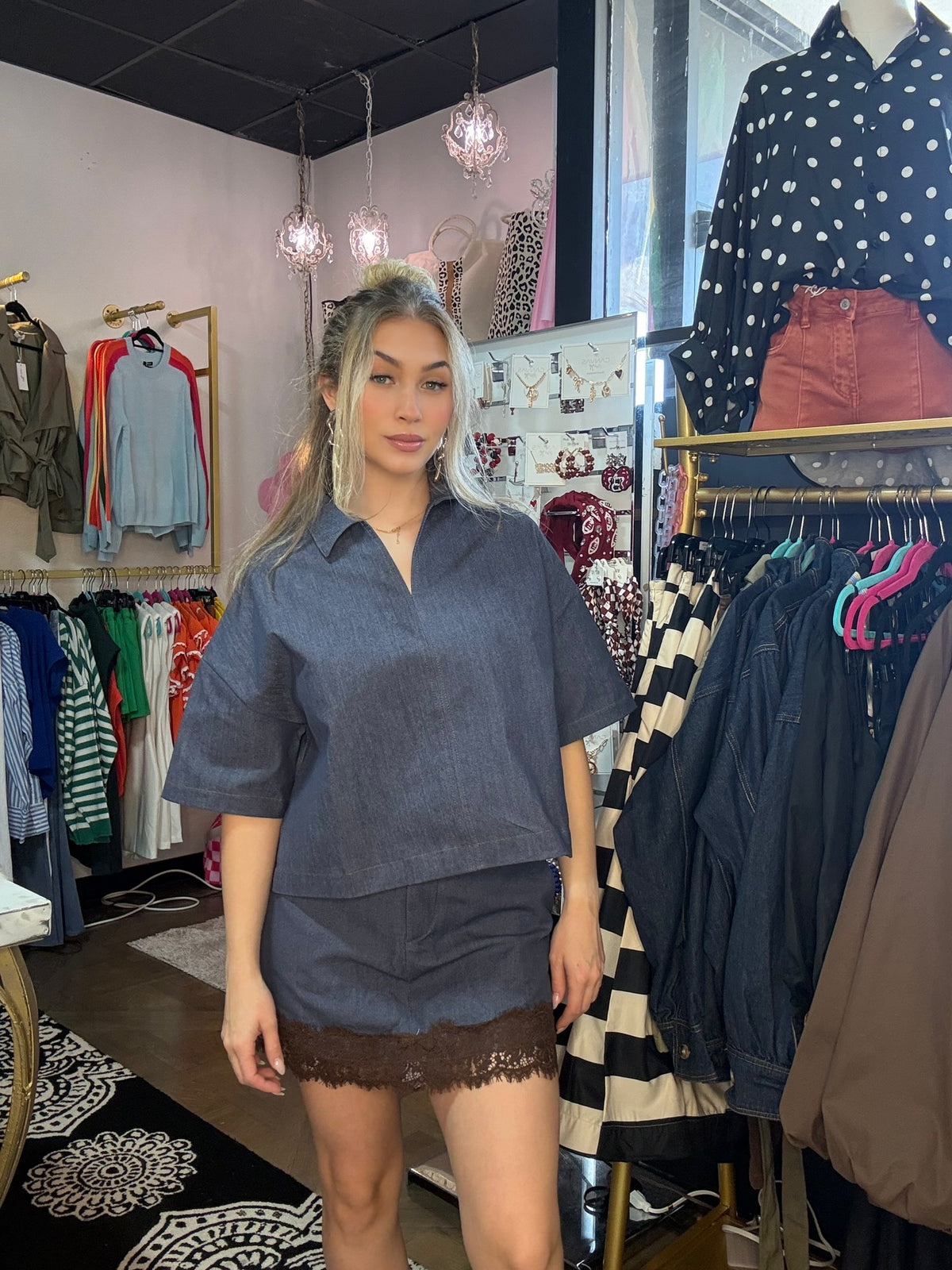 A woman with blonde-highlighted hair stands inside a clothing boutique wearing a dark slate blue short-sleeve collared shirt and matching shorts with a brown lace hem. Clothing racks and mannequins are visible in the background.