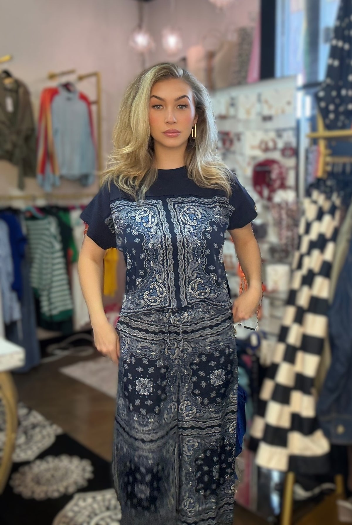 A woman modeling a short-sleeve maxi dress with a navy blue base and white paisley bandana print, standing inside a clothing boutique.