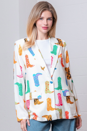 A woman wearing a white blazer with a colorful cowboy boot print, paired with a white top and blue jeans, standing against a white brick wall.