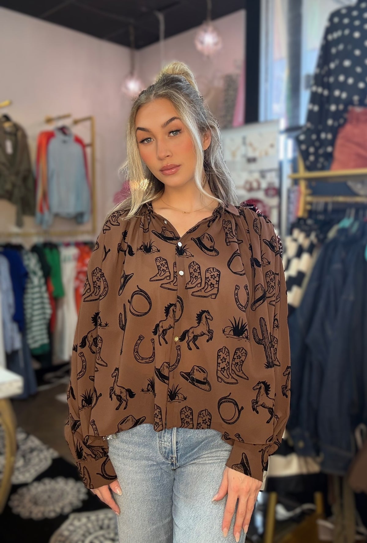 A woman wearing a brown long-sleeve button-up blouse with a black western-themed print of boots, horseshoes, cacti, and tumble stones, paired with light-wash blue jeans in a boutique setting.